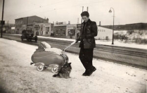Mann mit Kinderwagen und Hund auf schneebedecktem Gehweg, Läden im Hintergrund