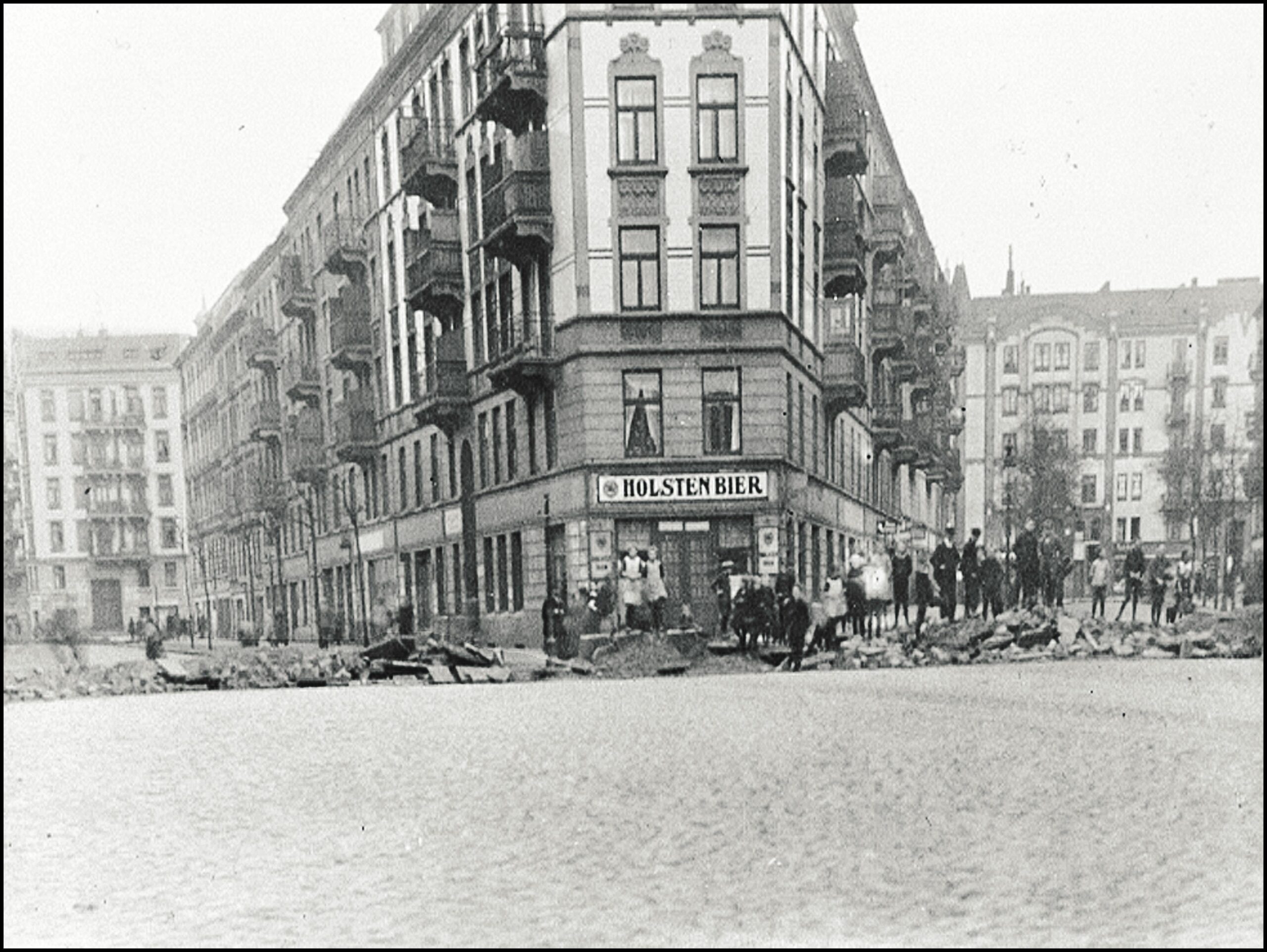 Menschen vor einer Barrikade mit Trümmern in einer Straße mit Eckkneipe Holsten Bier
