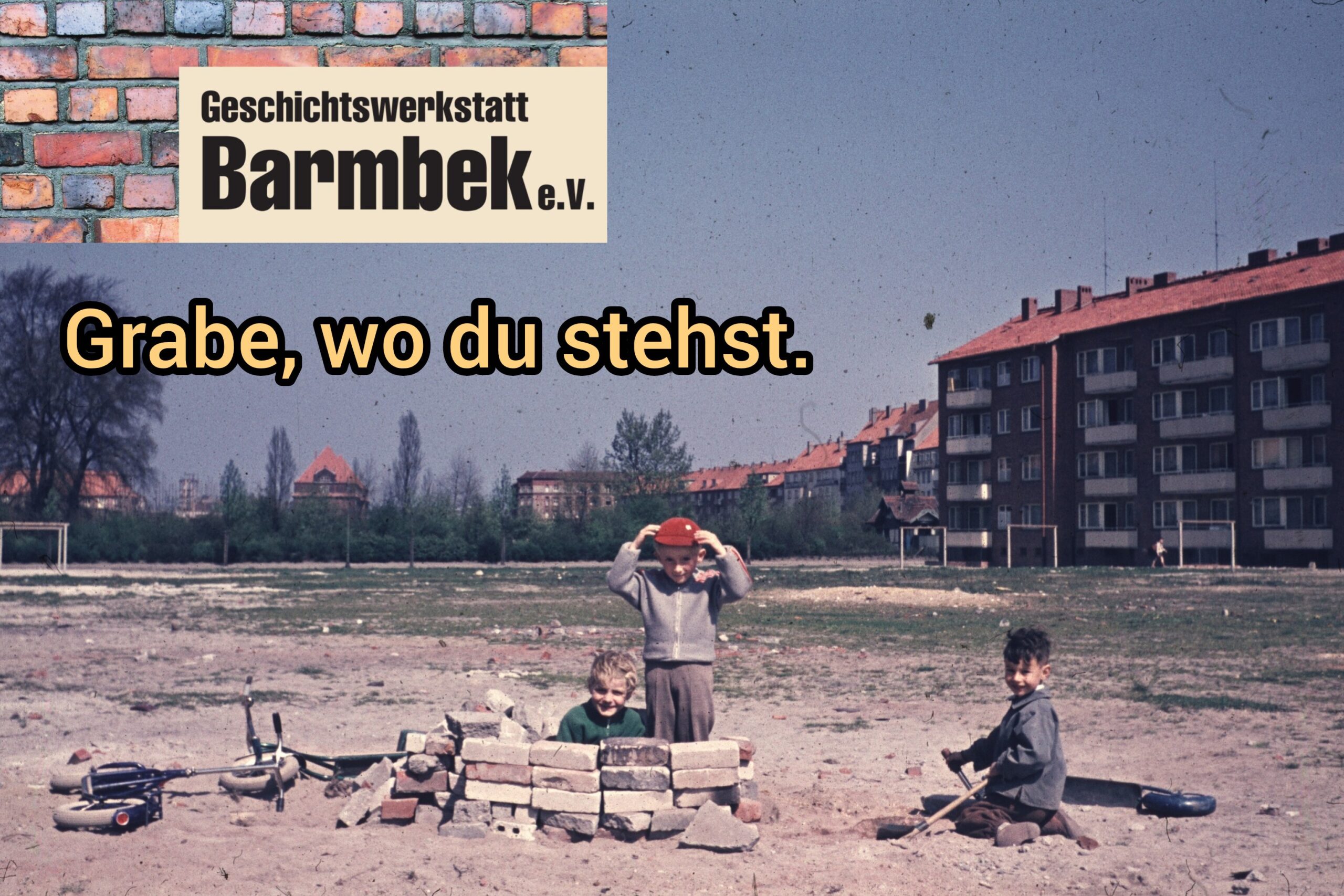 Drei Kinder spielen mit Ziegelsteinen auf einer Sandfläche, im Hintergrund Wohnhäuser.